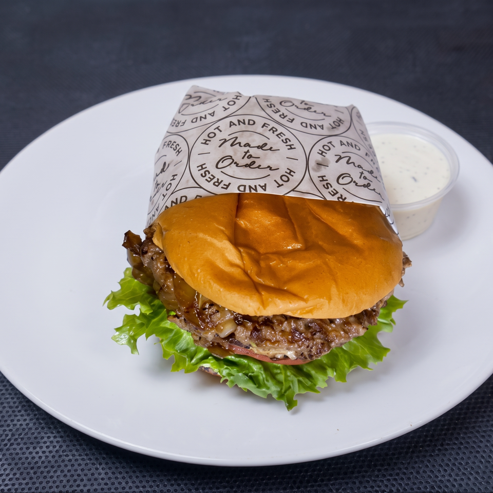 Juicy cheeseburger with lettuce, tomato, and caramelized onions on a white plate, accompanied by a cup of creamy dipping sauce.