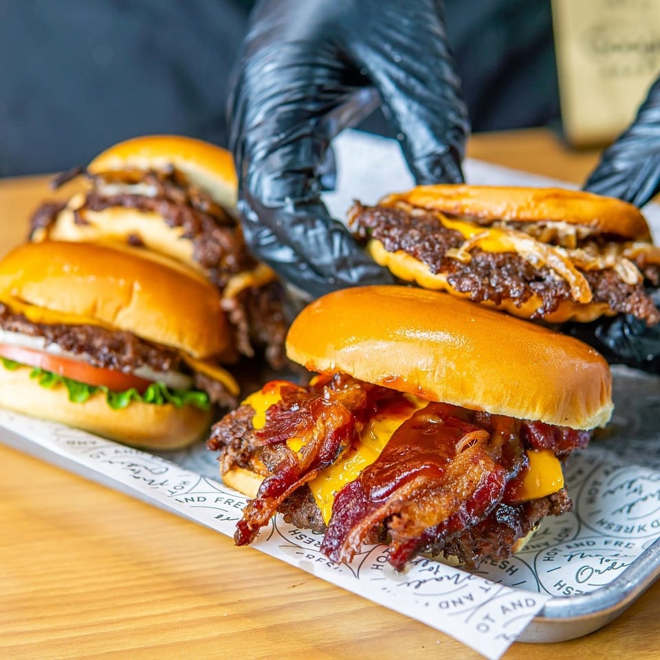 Four stacked cheeseburgers, including a bacon cheeseburger, sit on a paper-lined tray while gloved hands arrange them.